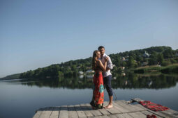 Couple embracing on a wooden dock overlooking a calm lake with green hills in the background.