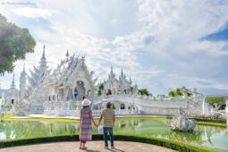 Couple holding hands facing a white ornate temple with a circular reflecting pool in front of them, under a blue sky.